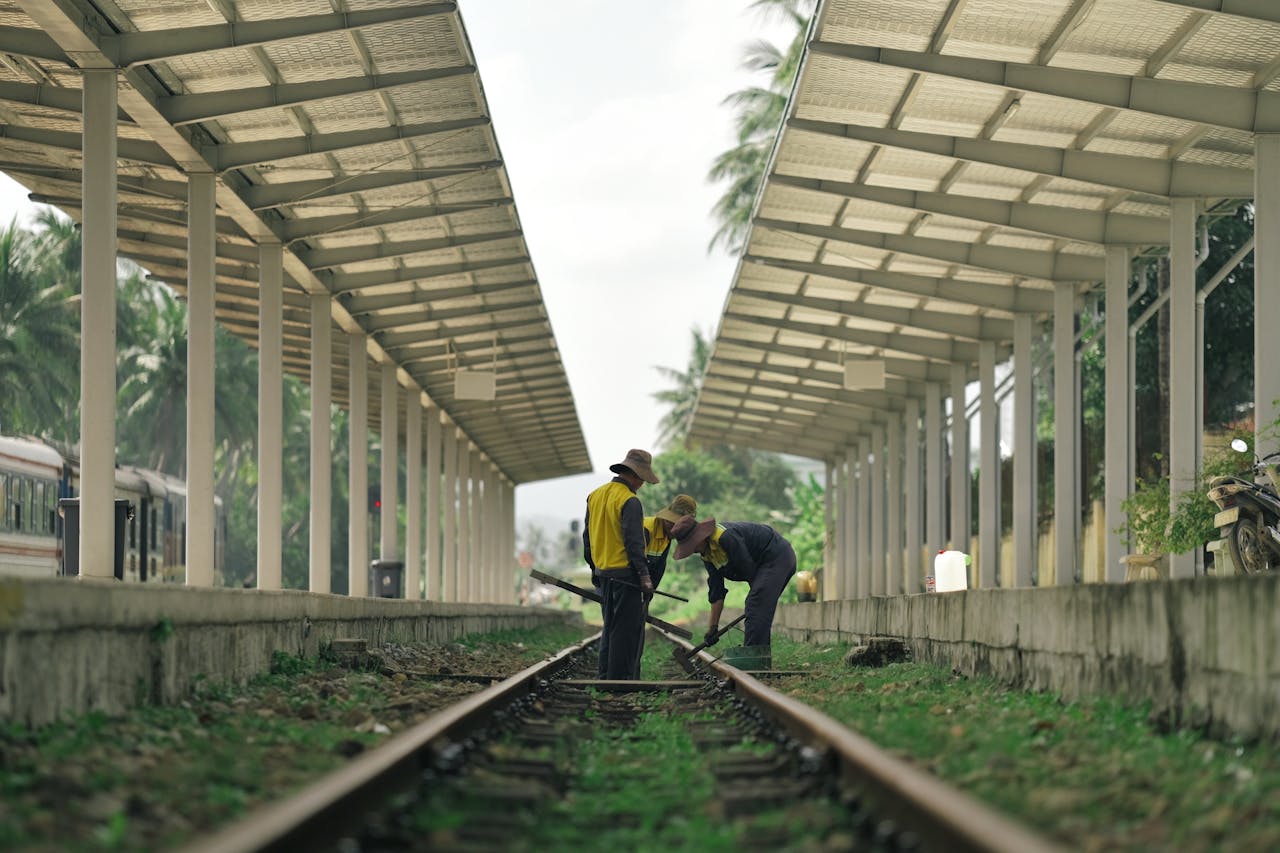 Workers fix railroad tracks at a rural, lush train station under a long canopy.